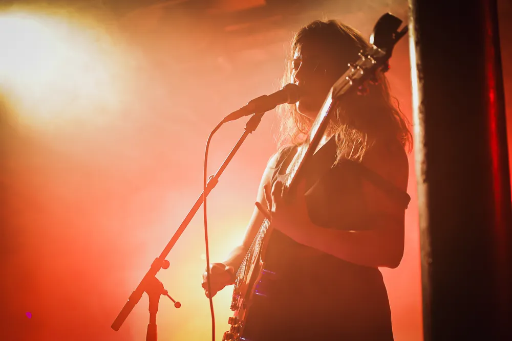 Silhouettenartige Aufnahme von Kee Avil mit Gitarre und Mikrofon im warmen B&uuml;hnenlicht.